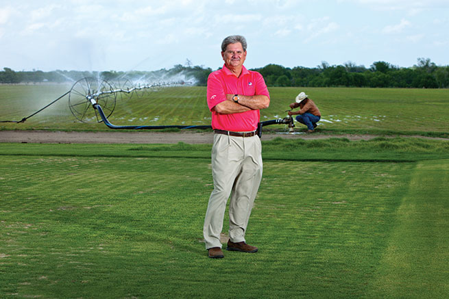 White man in golf shirt stands in front of irrigation sprinkler operated by Latino man in cowboy hat.